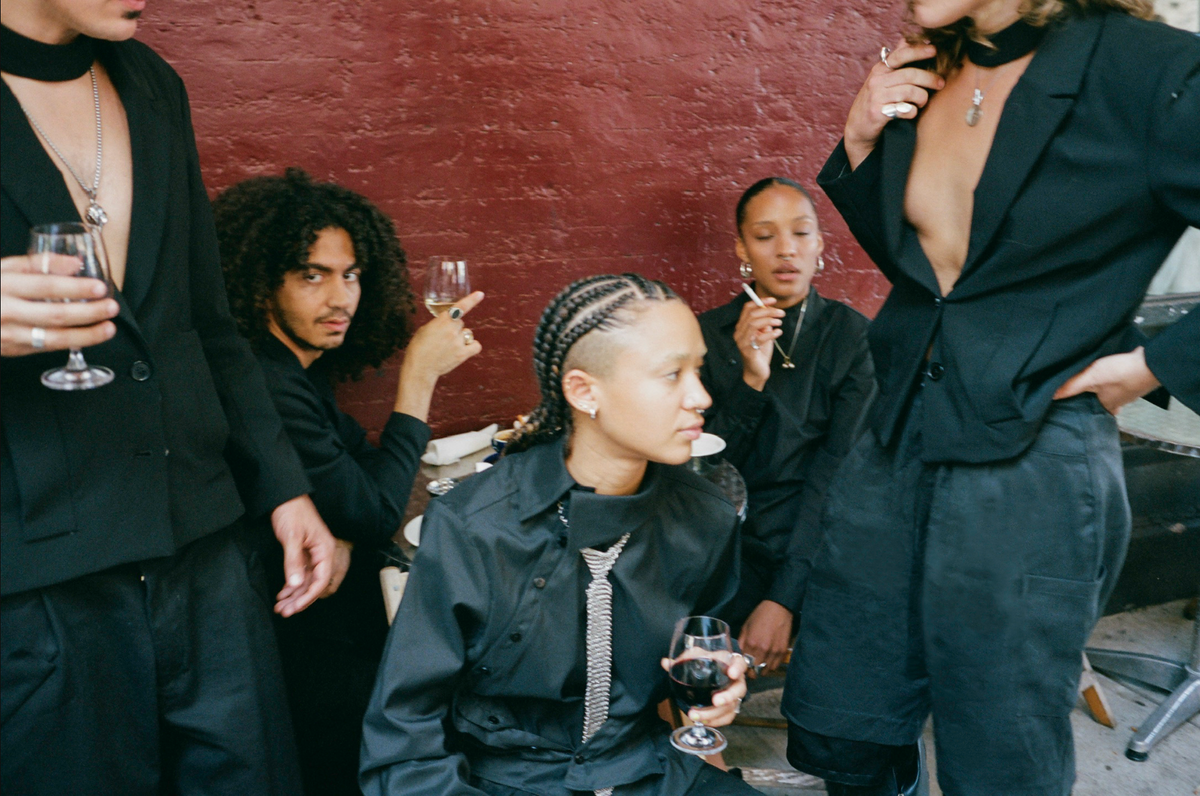 Group portrait in black tailoring against a red wall, wine glasses in hand