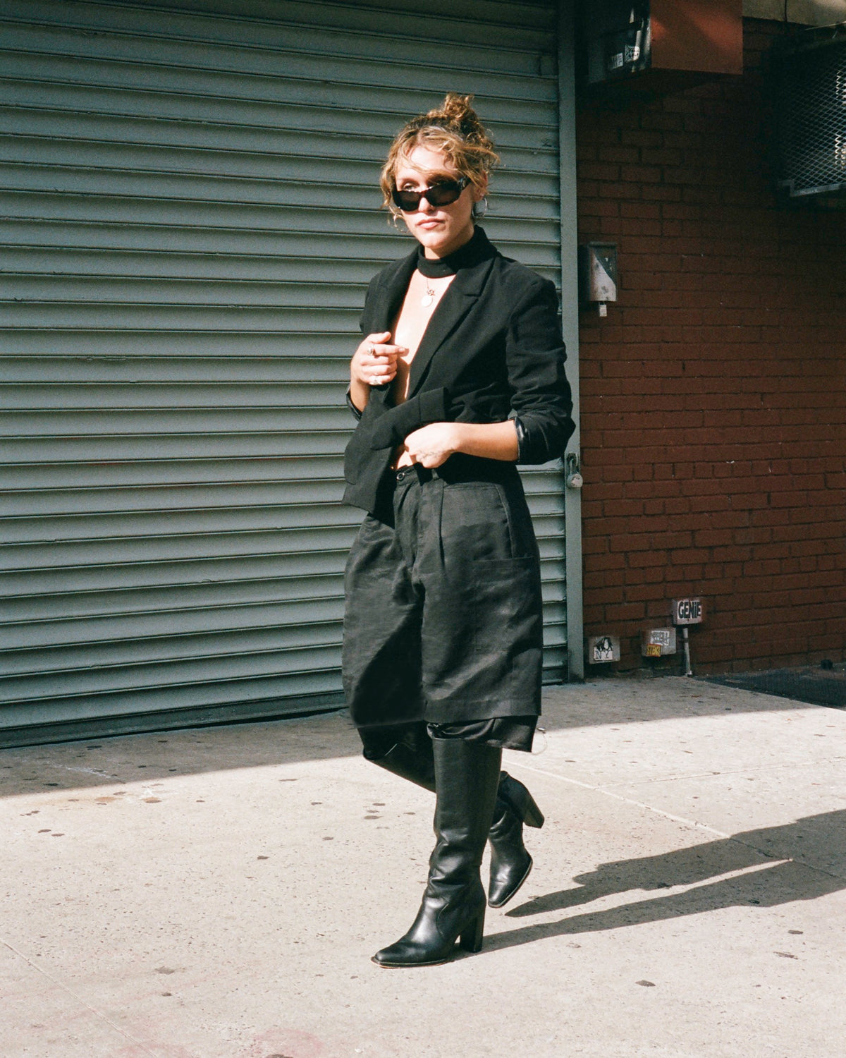 Figure in sunglasses, a black blazer and knee boots walking past a shutter