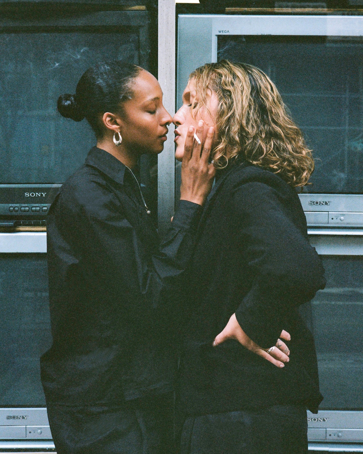 Two figures kissing in front of a wall of televisions